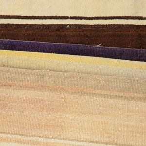 May include: Close-up of several folded woven rugs. The rugs display a variety of colors, including cream, brown, purple, yellow, and peach. The rugs have a striped pattern.