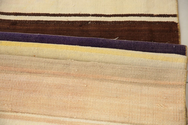 May include: Close-up of several folded woven rugs. The rugs display a variety of colors, including cream, brown, purple, yellow, and peach. The rugs have a striped pattern.