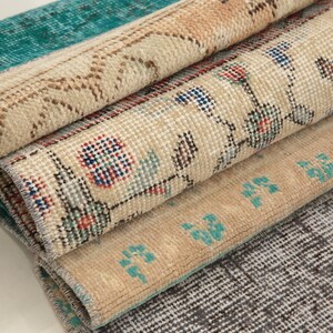 Könnte beinhalten: Ein Stapel von vier Vintage-Teppichen mit verschiedenen Mustern und Farben. Der oberste Teppich ist blaugrün und cremefarben, der zweite Teppich ist cremefarben mit einem Blumenmuster, der dritte Teppich ist cremefarben mit einem geometrischen Muster und der unterste Teppich ist grau mit einem gewebten Muster.