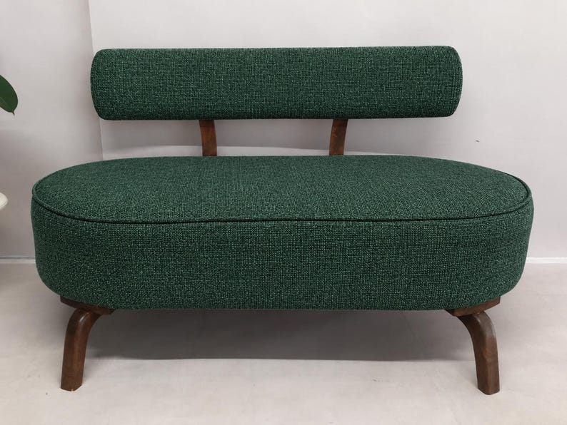 May include: A small loveseat upholstered in a textured dark green fabric. The piece features an oval-shaped seat, a rectangular backrest supported by two wooden struts, and four curved wooden legs with a dark brown finish.
