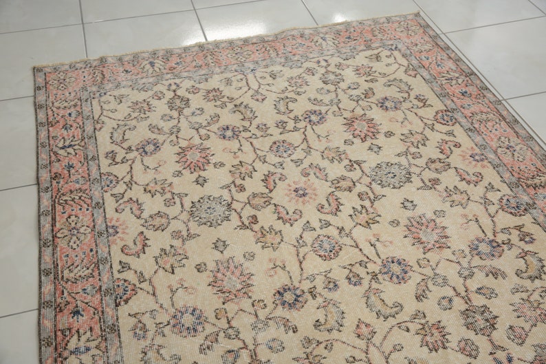 May include: A vintage rug with a cream background and a floral pattern in shades of pink, blue, and gray. The rug has a border with a repeating pattern of pink and gray.