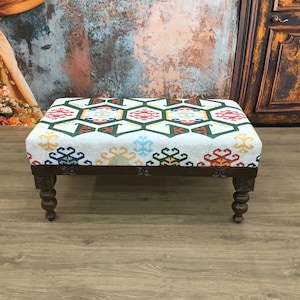 May include: A rectangular ottoman with a white fabric top featuring a colorful geometric pattern in red, green, blue, and yellow. The dark brown wooden legs are carved and have a turned design. The ottoman is a decorative furniture piece.