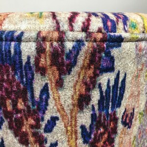 May include: Close-up of a round, cream-colored ottoman with a colorful floral pattern in shades of blue, purple, red, and orange. The fabric has a velvet texture.