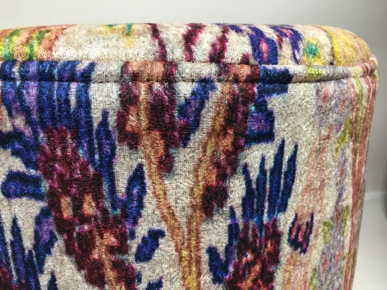 May include: Close-up of a round, cream-colored ottoman with a colorful floral pattern in shades of blue, purple, red, and orange. The fabric has a velvet texture.