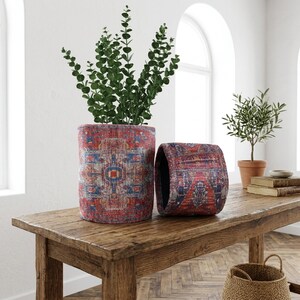 Puede incluir: Dos macetas cilíndricas con un diseño estampado en rojo, azul y blanco sobre una mesa de madera. Una maceta contiene una planta verde frondosa. La otra maceta está vacía. Un pequeño olivo en maceta de terracota al fondo.