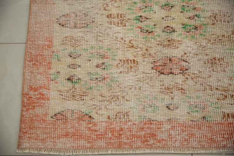 May include: A vintage rug with a cream background and a floral pattern in shades of brown, green, and pink. The rug has a peach-colored border and a fringe edge.