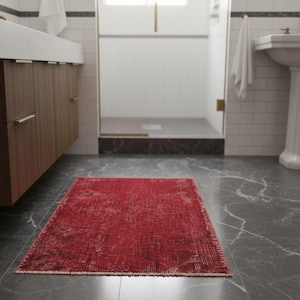 May include: A rectangular area rug featuring a deep red color palette with a distressed, faded pattern throughout. The rug has a low-pile texture and finished edges, positioned centrally on a dark gray marble floor within a bathroom setting.