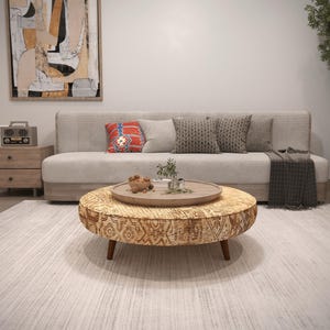 May include: A living room scene featuring a beige sofa with decorative pillows and a patterned throw. A round coffee table with a wooden tray and decor sits on a light-colored rug. An abstract painting hangs on the wall.