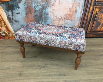 May include: A rectangular ottoman featuring a multi-colored fabric top with a detailed intricate pattern in shades of blue, red, and white. The seat is supported by four turned wooden legs with a smooth brown finish and a rounded bulbous design.