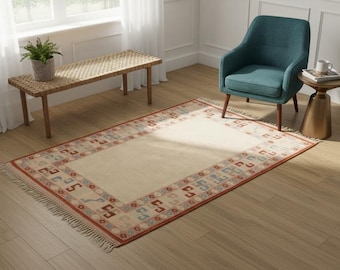 May include: A rectangular area rug featuring a solid cream-coloured centre and a decorative border with repeating geometric patterns in shades of red, blue, and tan. The rug has short fringe along the two shorter ends and a flat-woven texture.