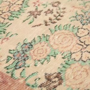 May include: Close-up of a beige rug with a floral pattern. The rug features a repeating design of pink and white flowers with green leaves. The rug has a subtle pink border.