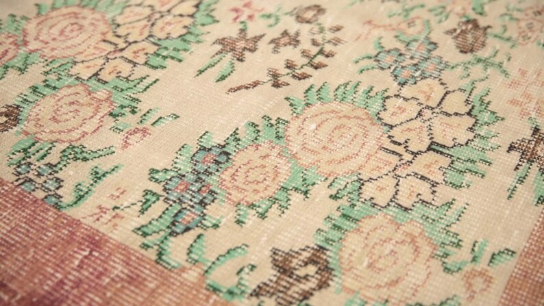 May include: Close-up of a beige rug with a floral pattern. The rug features a repeating design of pink and white flowers with green leaves. The rug has a subtle pink border.