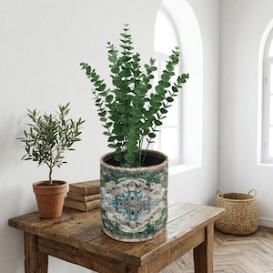 Op de afbeelding: Decoratieve binnenscène met een grote groene plant in een pot met patroon, een kleine olijfboom in pot en een rieten mand. De grote pot heeft een blauw, groen en wit ontwerp. De planten staan op een rustieke houten tafel.