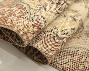 May include: A folded area rug featuring a woven pattern in shades of cream, beige, and brown. The textile displays intricate floral and geometric motifs with a visible grid-like weave structure, showing both the top surface and the underside edge.