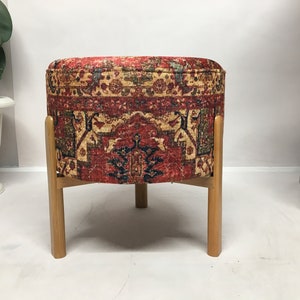 May include: A round ottoman with a red, blue and gold patterned fabric cover. The ottoman has four wooden legs.