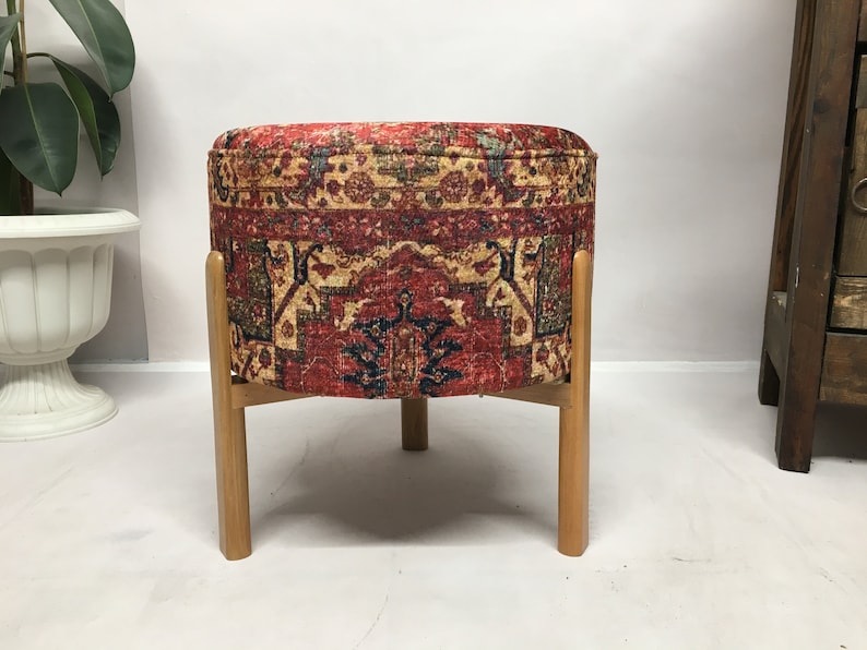 May include: A round ottoman with a red, blue and gold patterned fabric cover. The ottoman has four wooden legs.