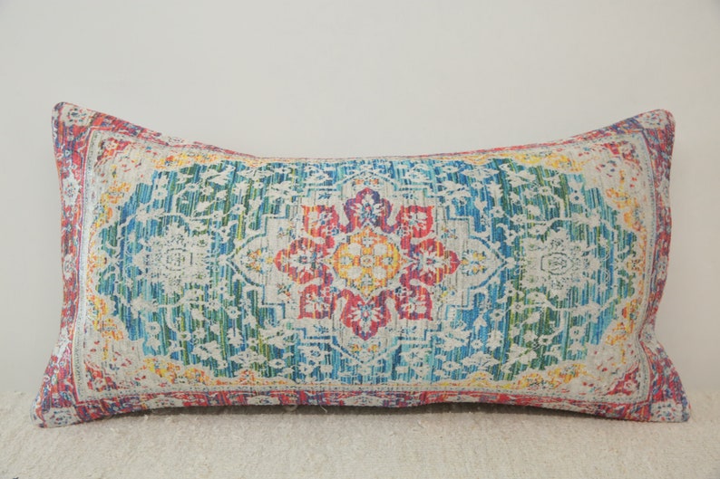 May include: A rectangular throw pillow with a colorful, intricate patterned design. The pattern features a central floral motif in shades of red, yellow, and blue, surrounded by a border of smaller floral designs. The pillow is made of a soft, textured fabric.
