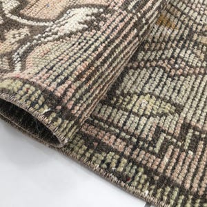 May include: A woven rug featuring a geometric pattern in shades of brown, muted pink, and pale green. The textile displays a dense, knotted pile construction with a visible edge binding and a folded corner revealing the underside texture.