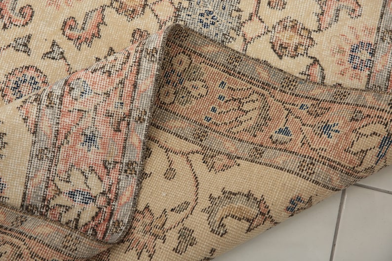May include: Close-up of the edge of a beige rug with a floral pattern. The rug has a fringe border and is woven with intricate details.