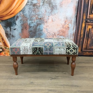 Puede incluir: Un otomano de madera con cuatro patas torneadas y una parte superior de patchwork hecha de tela colorida. El otomano está sentado en un suelo de madera frente a una pared con una superficie texturizada.