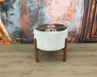 May include: A white, round ottoman with a colorful patterned top and four wooden legs. The ottoman is sitting on a wooden floor.