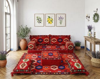 Puede incluir: Un juego de asientos de suelo rojo con un patrón geométrico, que incluye un sofá, una otomana y una alfombra. El conjunto está adornado con borlas y cojines decorativos. La habitación cuenta con estampados botánicos enmarcados, plantas en macetas y una mesa auxiliar de madera.