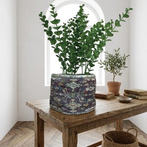 May include: A decorative planter with a patterned design, filled with vibrant green eucalyptus branches. The planter sits on a rustic wooden table, alongside a small potted olive tree and books. The room has a bright, airy feel.
