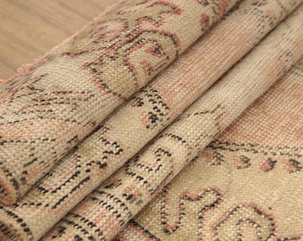 Può includere: Un tappeto piegato caratterizzato da un motivo intrecciato nei toni del beige, rosa tenue e marrone scuro. Il tessuto presenta un disegno geometrico e floreale ripetitivo con una superficie a pelo strutturato, che mostra l'intreccio complesso.
