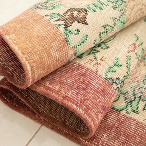 May include: Close-up of a folded, patterned rug with a floral design in shades of green, pink, and brown. The rug has a textured, woven surface.
