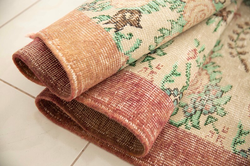 May include: Close-up of a folded, patterned rug with a floral design in shades of green, pink, and brown. The rug has a textured, woven surface.