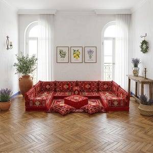 May include: A red sectional sofa with a patterned design, accompanied by matching pillows and a square ottoman. The room features framed botanical prints, a large terracotta vase, and a wooden side table with a vase of lavender.