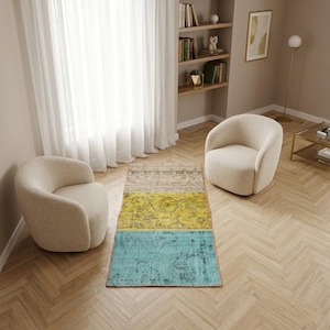 Könnte beinhalten: Rechteckiger Teppich mit drei Abschnitten: Beige, Gelb und Türkis. Der Teppich liegt auf einem Holzboden zwischen zwei cremefarbenen Sesseln. Der Raum hat eine neutrale Farbpalette mit Bücherregal und Fenster.