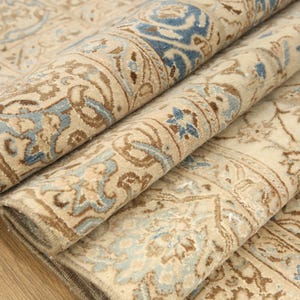 May include: A folded area rug featuring an intricate floral pattern in shades of cream, beige, and light blue. The textile displays a dense, woven design with symmetrical motifs and a detailed border, showcasing the texture of the pile and fabric.