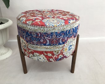 May include: Round ottoman with a patterned fabric top in red, blue, and white. The fabric has a floral design. The ottoman is supported by four wooden legs. A decorative piece for a living room or bedroom.