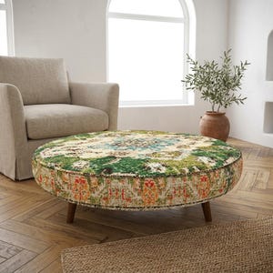 May include: A round ottoman with a colorful, patterned top featuring green, blue, and orange hues. The ottoman has a dark brown trim and sits on four wooden legs. A beige armchair is in the background.