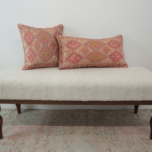 May include: A white upholstered bench with a wooden frame and turned legs. Two patterned pillows are placed on the bench, one square and one rectangular. The pillows have a pink and orange geometric design.