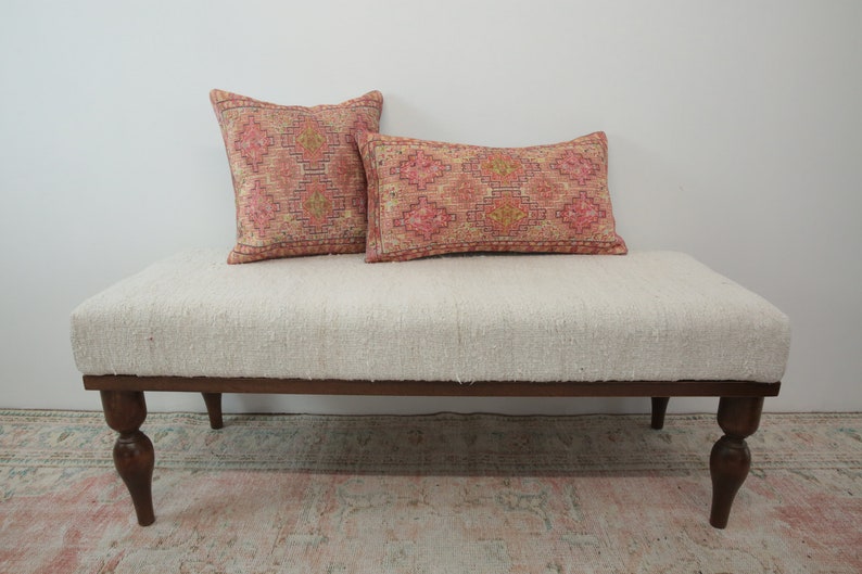 May include: A white upholstered bench with a wooden frame and turned legs. Two patterned pillows are placed on the bench, one square and one rectangular. The pillows have a pink and orange geometric design.
