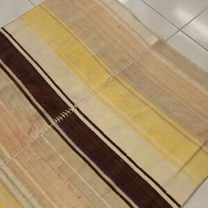 May include: A rectangular rug with a patchwork design. The rug features alternating stripes of brown, yellow, and beige. The rug is made of woven fabric and has a textured appearance. The edges are finished with a fringe.