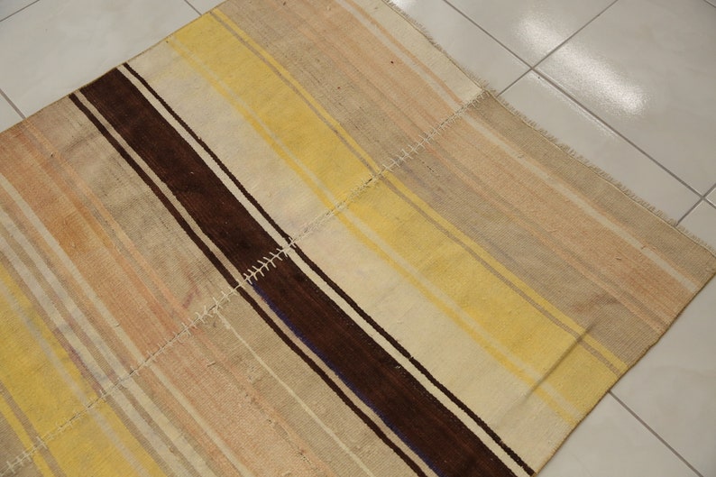 May include: A rectangular rug with a patchwork design. The rug features alternating stripes of brown, yellow, and beige. The rug is made of woven fabric and has a textured appearance. The edges are finished with a fringe.