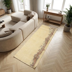 Puede incluir: Una alfombra rectangular de color crema con un borde detallado de patrones florales marrones y rojos. La alfombra está colocada sobre un suelo de madera clara, junto a un sofá seccional beige. La habitación está bien iluminada.