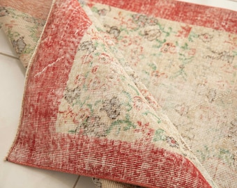 May include: A woven area rug with a folded corner revealing the underside. The rug features a cream-coloured base with a floral pattern in shades of red, green, and black, framed by a wide, solid red border along the edges of the fabric.