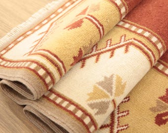 May include: A folded area rug featuring a geometric pattern in shades of rust red, cream, and tan. The rug has a thick, textured pile with a visible fringe along the edge and a repeating motif of stylized leaf-like shapes and linear borders.