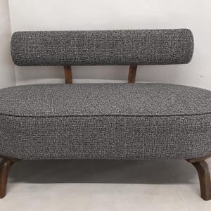 May include: A small loveseat featuring a rounded, oval-shaped seat upholstered in a textured grey fabric. It includes a matching rectangular backrest supported by two wooden posts and stands on four curved, dark brown wooden legs.