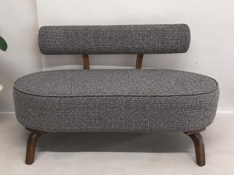 May include: A small loveseat featuring a rounded, oval-shaped seat upholstered in a textured grey fabric. It includes a matching rectangular backrest supported by two wooden posts and stands on four curved, dark brown wooden legs.