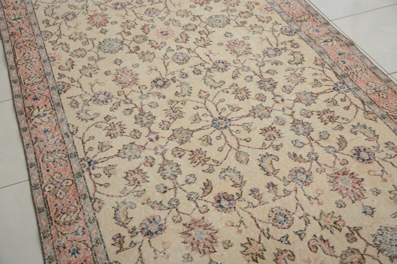 May include: A vintage cream-colored rug with a floral pattern in shades of pink, blue, and brown. The rug has a border with a repeating pattern of pink and brown.