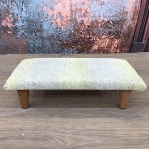May include: A small, rectangular footstool with a light gray and yellow striped fabric top and wooden legs.