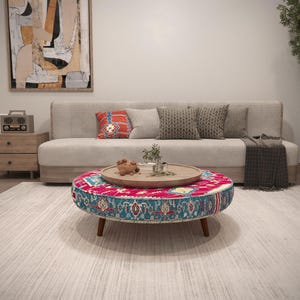 May include: A living room scene featuring a beige sofa with decorative pillows and a patterned throw blanket. A round, low coffee table with a colorful, patterned top and wooden legs sits on a light-colored rug. A wooden tray with decorative items rests on the table.