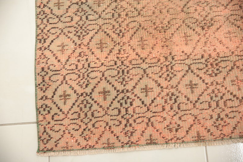 May include: Close-up of a pink and brown patterned rug with a fringe border. The rug has a geometric design with a repeating pattern of diamonds and crosses.