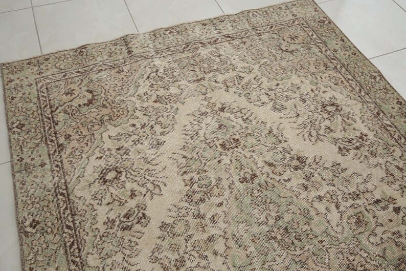K&ouml;nnte beinhalten: Ein Vintage-Teppich mit beigefarbenem Hintergrund und einem floralen Muster in Braun- und Gr&uuml;nt&ouml;nen.
