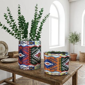 May include: Two cylindrical fabric planters with colorful geometric patterns. One planter is larger and holds green leafy stems. The smaller planter has a similar design with orange, blue, and green hues. Both sit on a wooden table.